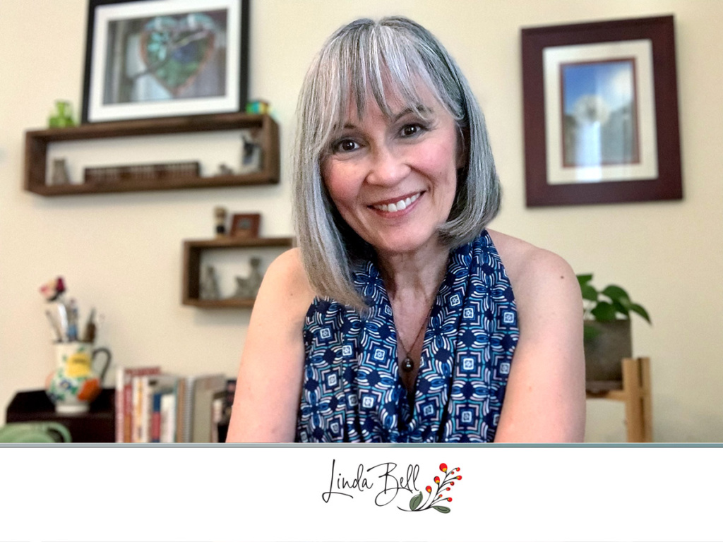 Portrait of Linda Bell smiling in her home studio, wearing a blue patterned scarf and sitting in front of wall shelves and framed artwork.